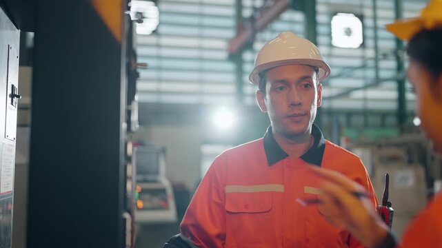 Factory Floor Discussion: Two industrial workers in hard hats and safety vests engage in conversation on a brightly lit factory floor, discussing a project.