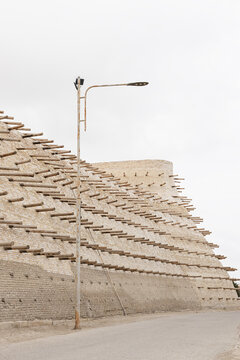 Ark Fortress wall in Bukhara with rows of protruding wooden beams rising against an overcast sky. Massive brick fortification with a modern street lamp in the foreground.