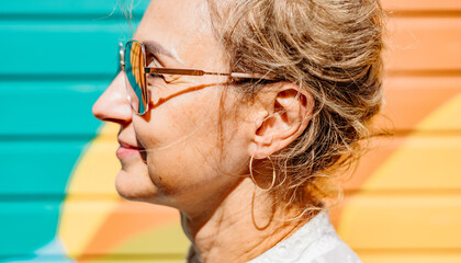 Profile of a middle-aged Caucasian woman with short, curly blond hair and sunglasses. She stands against a colorful mural background. © Alina