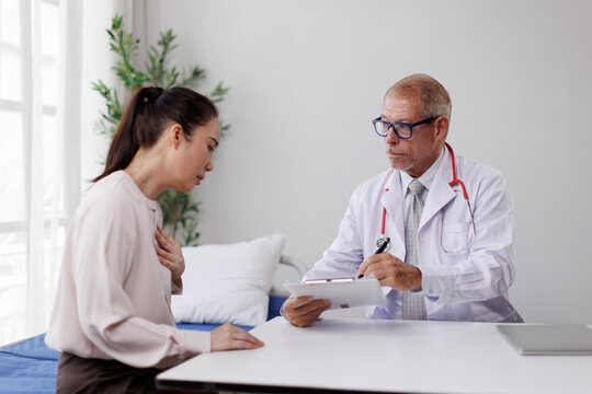 Senior doctor interviewing a distressed young female patient holding her chest, listening attentively while taking notes on a clipboard during a medical consultation in a clinic office