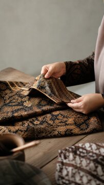 Close-Up of Hands Holding and Examining Luxurious Indonesian Batik Fabric with Intricate Gold and Navy Floral Pattern on Rustic Wooden Table, Earthy Heritage Textile Craft Concept