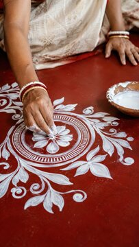 Authentic Indian Woman in White Sari Drawing Traditional White Floral Alpona Rangoli Pattern on Red Floor with Rice Paste, Earthy Warm Cultural Heritage Ritual and Folk Art Concept