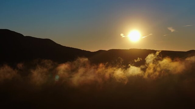 Aerial drone view of sunrise through glowing golden fog and low cloud layer over mountain ridge silhouette, with clear blue sky above and warm lens flare