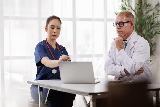 Female nurse and male doctor in scrubs and white coat review a laptop together in a clinical office, focused on teamwork, digital records and patient care planning