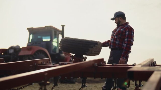 Technician repairing wheel of Agricultural machinery, hard work in rural area. Portrait of male farmworker in field, tractor driver checking wheel of tractor or harvester combine in farmland in summer