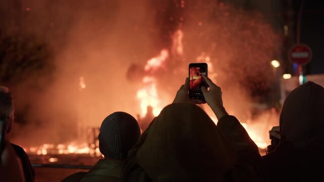 A person films a burning falla structure during Las Fallas La Crema in Valencia, orange flames filling the scene