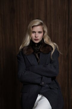 Blonde woman posing in dark grey winter coat with fur collar, holding black leather gloves near face in studio setting. fashion, winter, portrait, elegance, style
