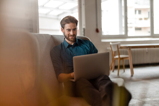 Portrait of young freelancer sitting on couch in a loft using laptop