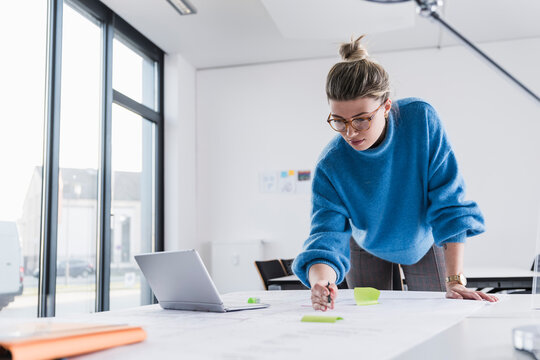 Young woman with laptop working on plan at desk in office