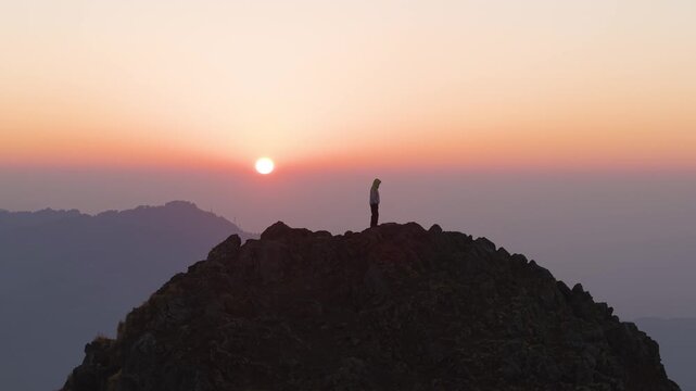 4K aerial view of a young traveler standing on a rocky cliff overlooking the Himalayan sunrise in Uttarakhand, India.