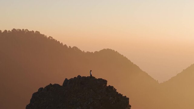 4K aerial shot of a boy standing on a cliff edge overlooking sunrise skies in the Himalayas of India.