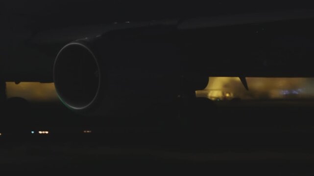 Jet engine intake viewed from below at night, circular nacelle rim in darkness, warm yellow airport terminal light glowing in background, minimal exposure
