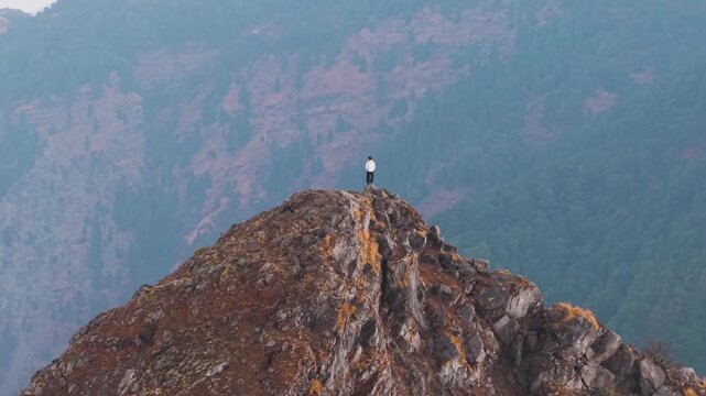 4K aerial 360 view of a boy standing atop a rugged mountain surrounded by layered Himalayan ranges in India.