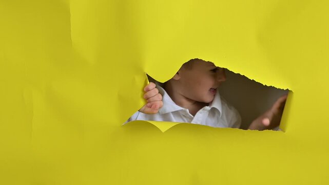 A mischievous boy peeks out through a hole in a yellow cardboard background. 