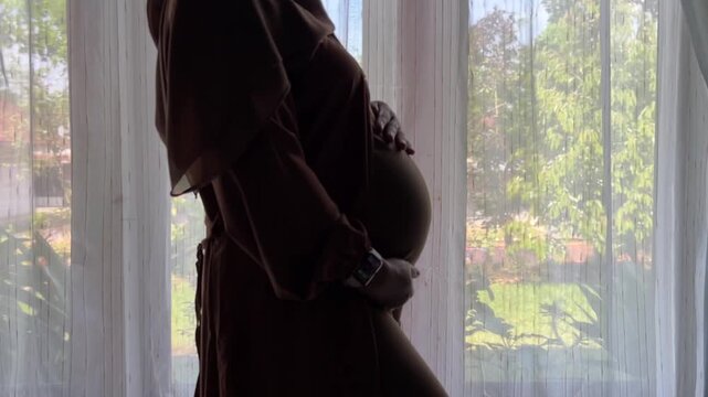 A pregnant Indonesian woman wearing a hijab stands in front of a window holding her stomach and her unborn baby. She and her first child kiss their mother's stomach.