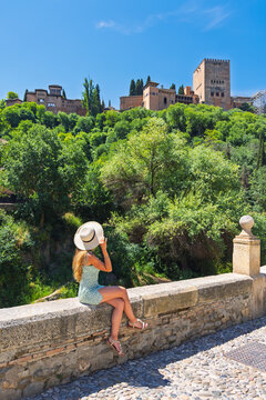 Woman tourist in dress and hat sitting on wall, looking at Ancient arabic fortress Alhambra- Granada in Spain
