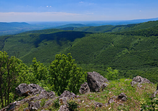 Summer landscape from Hegyes-kő featuring forested valleys, rocky foreground and dramatic cloud shadow over the green hills of B&uuml;kk National Park.
