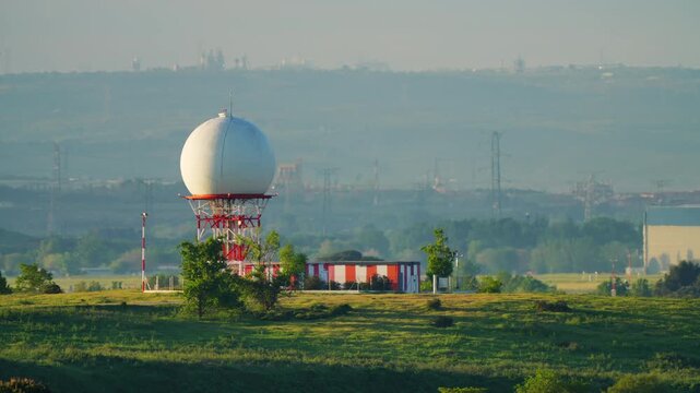 White spherical weather radar dome on red and white steel lattice tower in flat green airfield, misty hilly landscape and power line in background, warm sunrise light