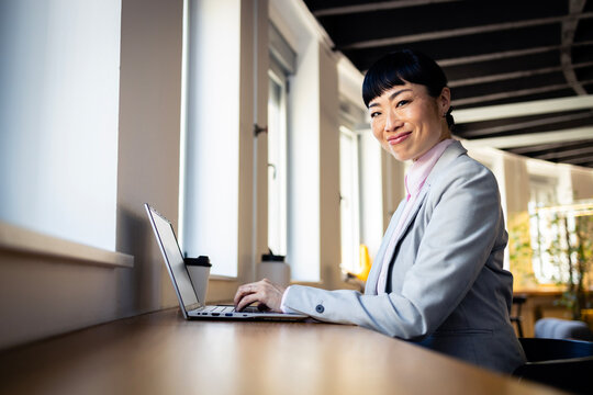 Asian businesswoman working on laptop in modern office, smiling confidently and looking at camera
