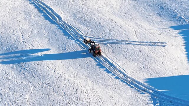Drone video captures a red tractor hauling equipment across an expansive, untouched snowy field under crisp morning light, casting long shadows over textured winter terrain. (198 characters)