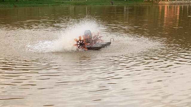 Mechanical paddlewheel aerator splashing muddy pond water, creating ripples and increasing oxygenation in a landscaped park