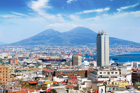 Panoramic skyline of Naples with Mount Vesuvius above busy harbor under blue sky. Urban districts, port infrastructure, Mediterranean coastline define Naples cityscape in Italy during summer