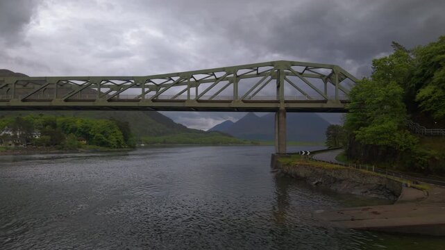Drone tracking shot alongside the Ballachulish Bridge spanning Loch Linne, with the moody, cloud-covered peaks of Glencoe visible in the background.