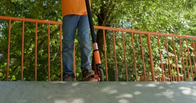 Boy stands with orange scooter near red railing in park area. Child holds handlebar upright keeping front wheel steady before starting another ride soon
