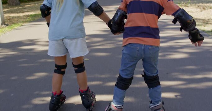 Brother and sister roll away together on inline rollerblades while holding hands. Children angle skates outward keeping shared balance during slow park practice