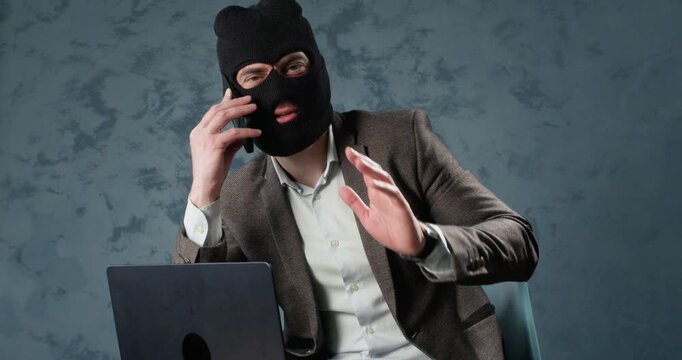 Masked scammer talks on smartphone near laptop during online extortion. Man gesticulates with free hand while continuing deceptive phone fraud conversation slow motion
