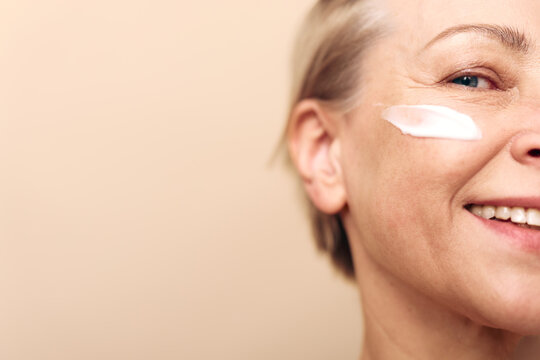 Close-up of smiling middle aged woman with cream on cheek against beige background. Concept of skincare, anti aging, self care, and natural beauty in wellness routine.