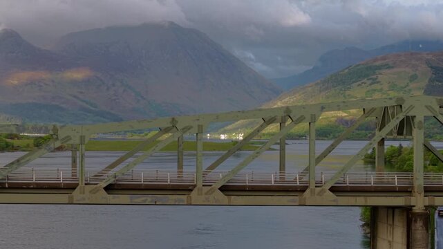 Telephoto drone tracking shot of the Ballachulish Bridge steel truss, with the dramatic, light-dappled mountains of Glencoe compressed in the background.