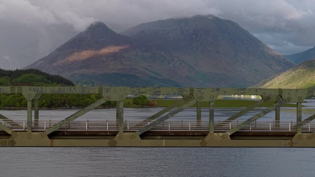 Telephoto drone tracking shot compressing the green steel truss of the Ballachulish Bridge against the dramatic, cloud-capped mountains of Glencoe.