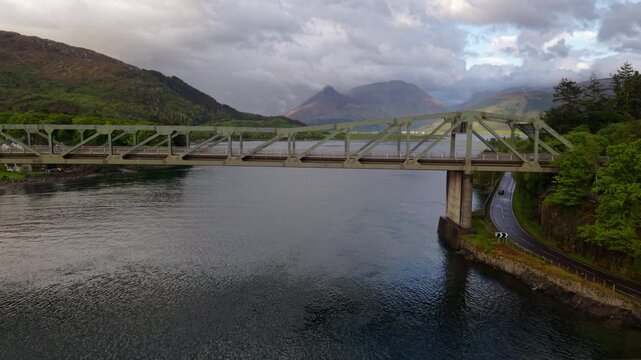 Drone flies slowly toward Ballachulish Bridge with stunning light on Glencoe seen in the background.