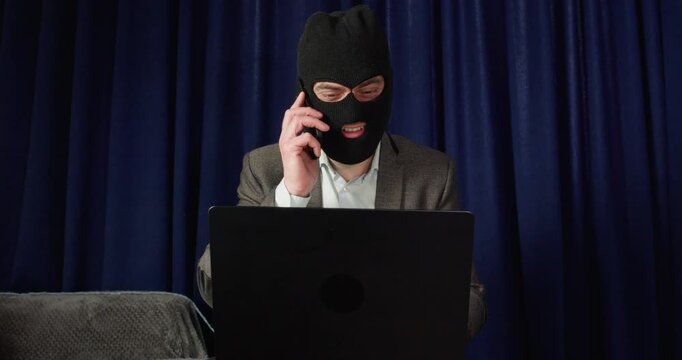 Masked scammer calls victim from behind laptop during money extortion. Man smiles while speaking on smartphone controlling digital fraud process at computer