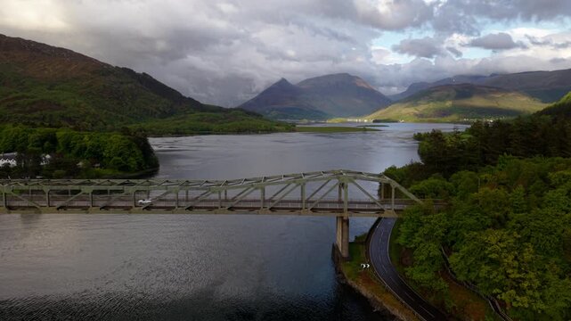 Shallow drone orbit around the Ballachulish Bridge, showcasing its steel truss structure against the expansive, sun-dappled mountains of Glencoe.