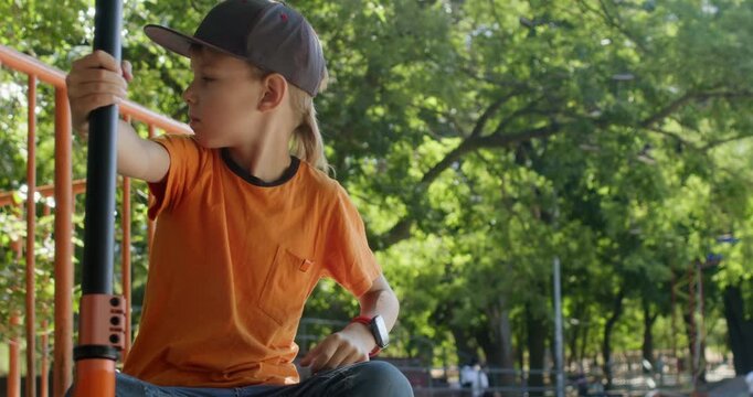 Junior schoolboy leans on scooter touching chin while sitting near park railing. Child holds handlebar while pausing in thought before returning to ride across pathway