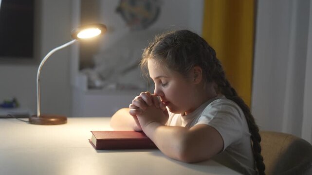 Young girl sitting at table with her hands on her head. Religious girl praying to a child. Religious book of prayer with hands on a table. A young girl is at a table with her hands on lifestyle her.
