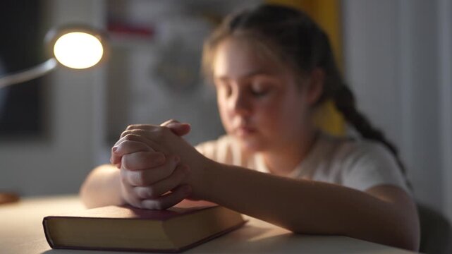Young girl sitting at table with her hands on her head. Religious girl praying to a lifestyle child. Religious book of prayer with hands on a table. A young girl is at a table with her hands on her.
