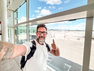 Man taking a cheerful selfie at an airport terminal with backpack, giving thumbs up while waiting near the gate with runway view conveying travel excitement and confident adventure © simona
