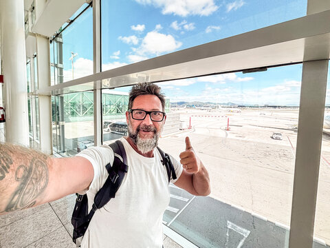 Man taking a cheerful selfie at an airport terminal with backpack, giving thumbs up while waiting near the gate with runway view conveying travel excitement and confident adventure