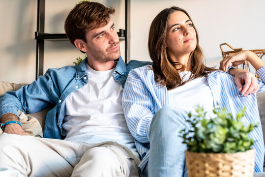 Young couple relaxing on a sofa at home, sharing a quiet moment together and looking thoughtfully into the distance, conveying comfort, intimacy, and hopeful planning for the future.