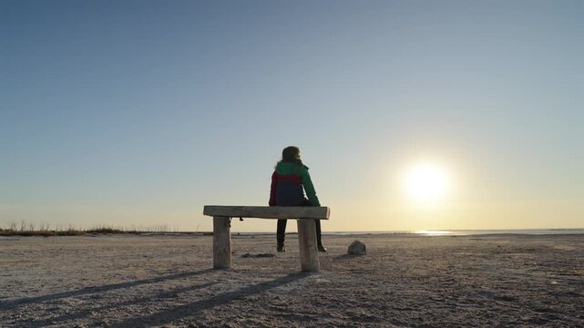 Child seated on bench in empty barren terrain under low sun with long shadows emphasizing loneliness vulnerability and isolation in minimal environment, static wide shot
