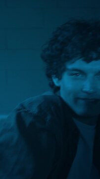 Vertical cinematic medium shot of young male performer performing expressive freestyle and smiling at camera with confidence illuminated by blue neon lighting in creative space, story format