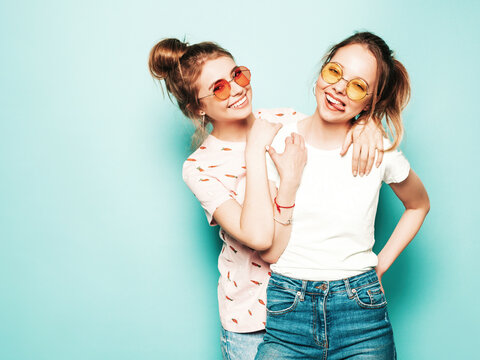 Two young beautiful blond smiling hipster in trendy summer hipster jeans clothes. Sexy carefree women posing near blue wall. Trendy and positive models having fun in sunglasses