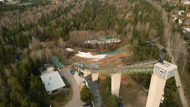 Aerial perspective circling around an old, abandoned ski jump tower and ramp, showcasing the concrete structure nestled within a vast pine forest with patches of melting snow