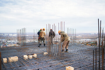 A team of construction workers in hard hats walk across a complex steel rebar grid with vertical dowels on a high-rise building slab overlooking a wide cityscape. © Aigul Sabirova