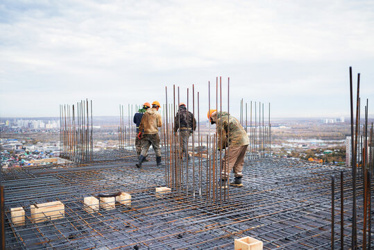 A team of construction workers in hard hats walk across a complex steel rebar grid with vertical dowels on a high-rise building slab overlooking a wide cityscape.