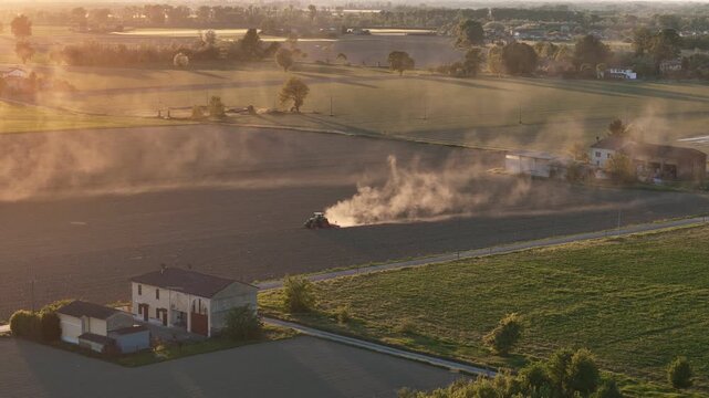 Tractor performing spring soil preparation near Strada Padana Inferiore in Province of Piacenza Emilia Romagna Italy as drought conditions create drifting dust plumes at sunset, drone tracking wide
