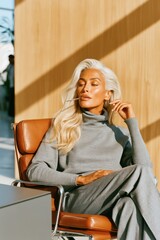 Obraz premium Confident mature woman in gray turtleneck and trousers relaxing in a modern office chair in sunlight